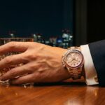 虹色のベゼルとダイヤモンドが輝くロレックス コスモグラフ デイトナ レインボー (Ref. 116595RBOW) を着用した、高級スーツ姿の日本人男性の手元。背景は東京の煌びやかな夜景。定価が存在しない幻の時計の圧倒的な資産価値と成功者のステータスを象徴する様子。